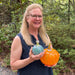 A woman with blonde hair and glasses holds two decorative glass pumpkins outdoors. One pumpkin is a vibrant orange, and the other is a textured light blue-green.