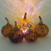 Five decorative glass pumpkins with amber and purple marbled patterns, arranged on a white surface with warm light projected behind them.