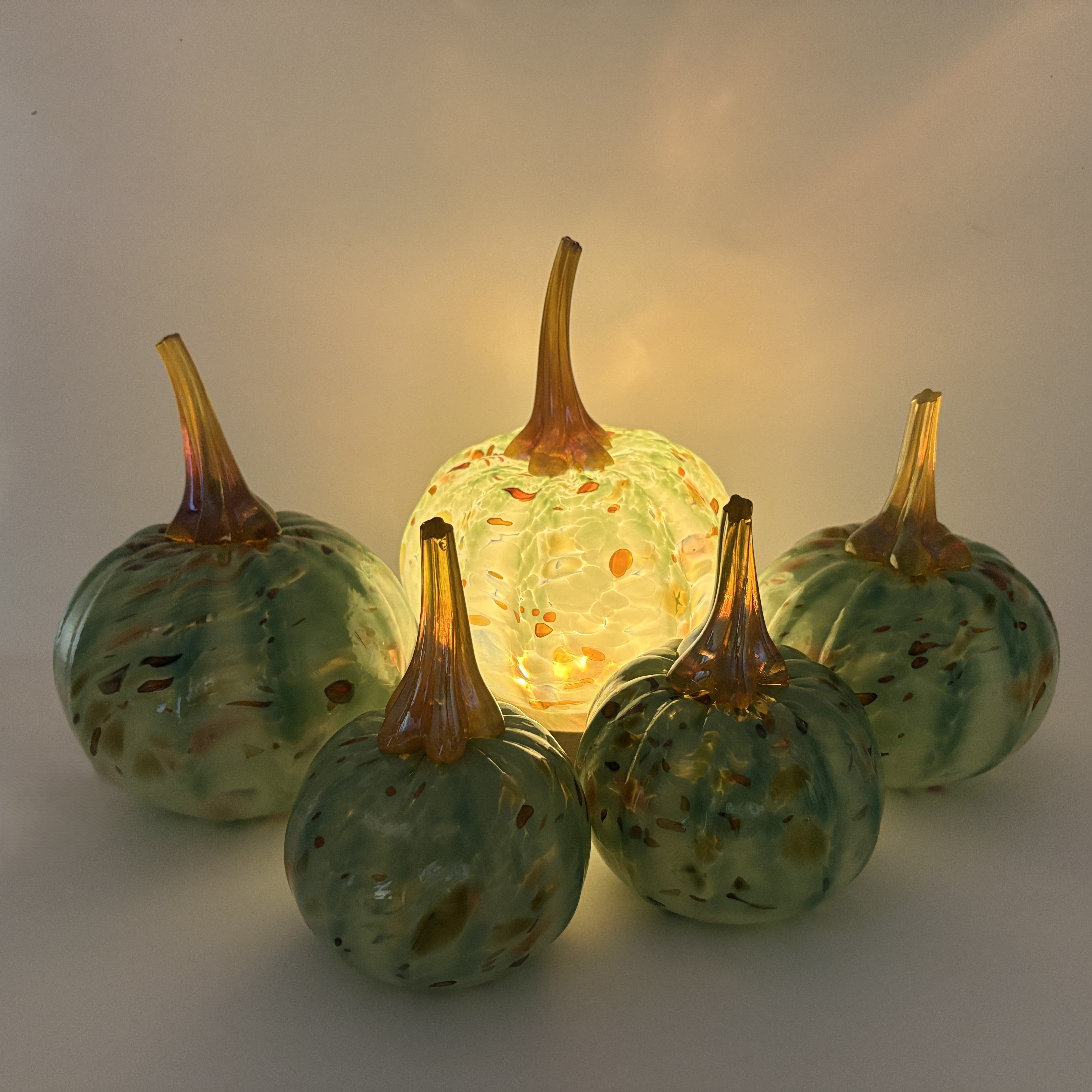 Four decorative glass pumpkins arranged on a light neutral surface, with one central pumpkin illuminated from within to cast a warm glow. The pumpkins have marbled green and blue glass bodies with amber-brown stems and small orange flecks.
