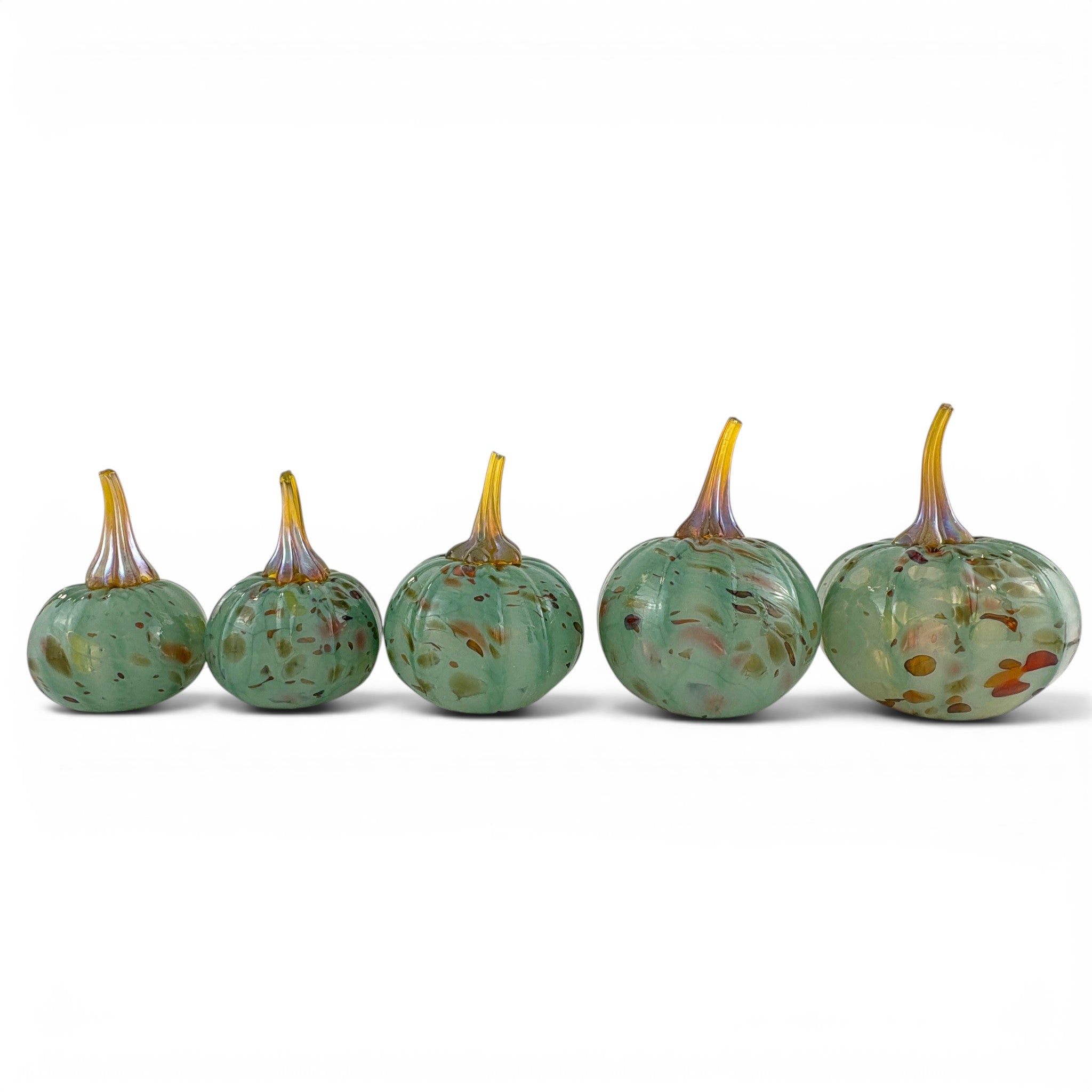 Five small decorative glass pumpkins in a row, made of speckled light green glass with yellow and orange accents on the stems, set against a plain white background.