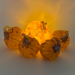 Five decorative glass pumpkins arranged together, with one larger pumpkin in the center glowing warm yellow-orange light. The pumpkins are translucent amber glass with darker brown stem details and are photographed against a softly lit neutral background.