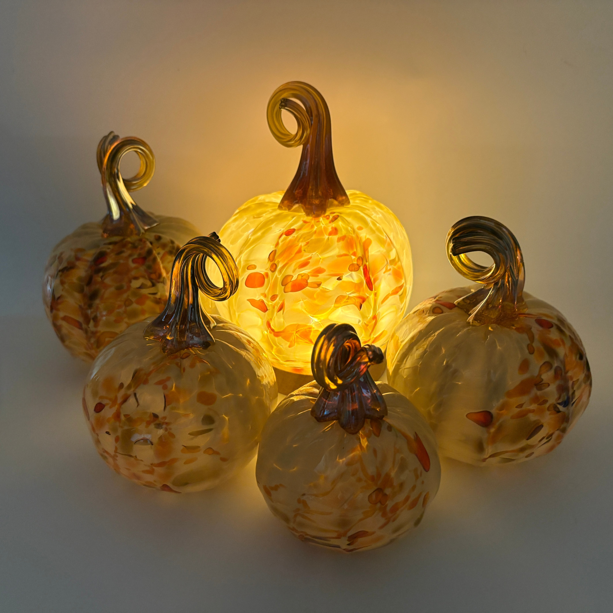 Four decorative glass pumpkins arranged together, glowing warmly from an internal light. The pumpkins are translucent amber and yellow with orange speckles and metallic gold-toned stems, set against a softly lit neutral background.