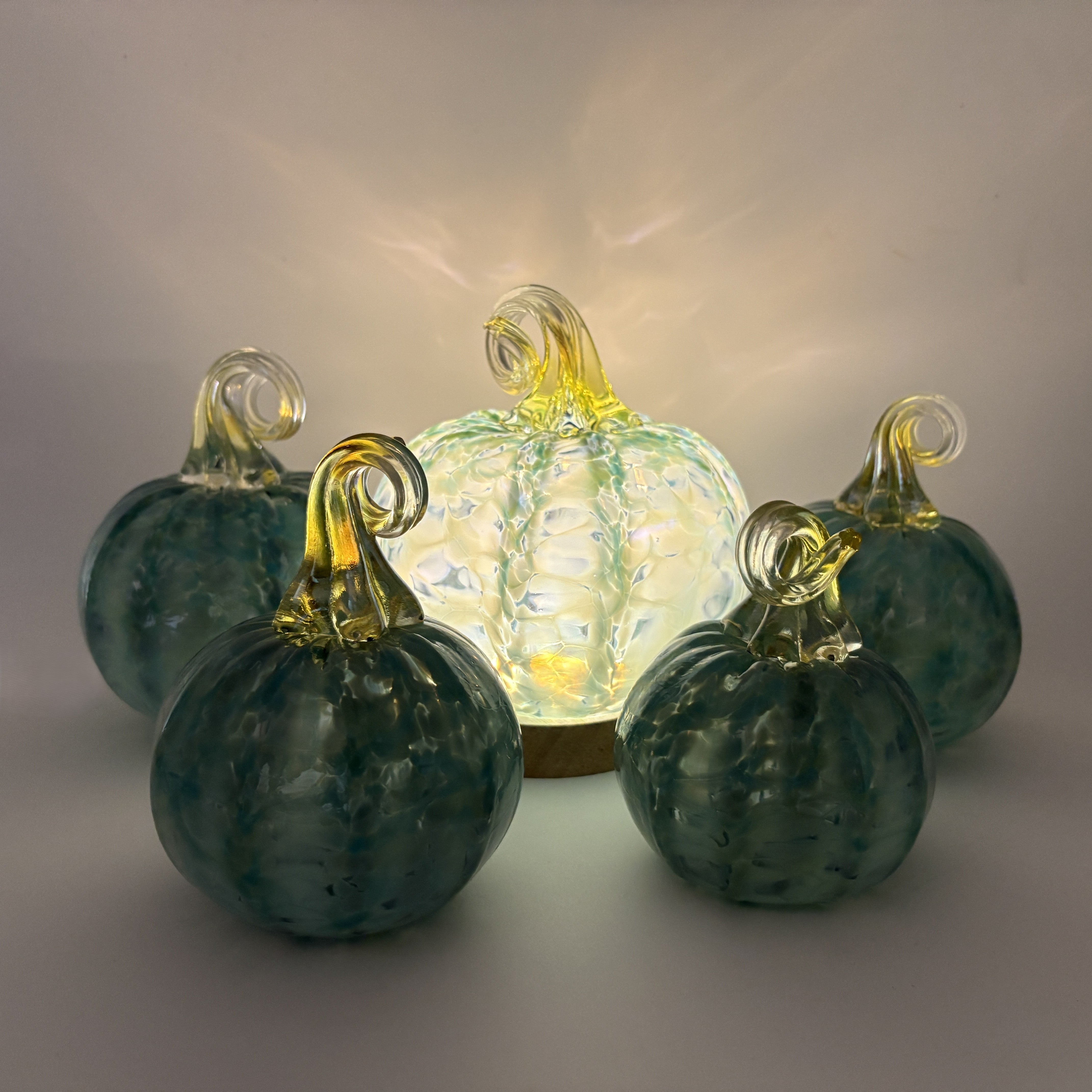 Four decorative glass pumpkins arranged on a light gray background, with one larger glowing pumpkin in the center and three darker teal pumpkins in front and behind. The pumpkins have ribbed textured bodies and translucent yellow-green stems.
