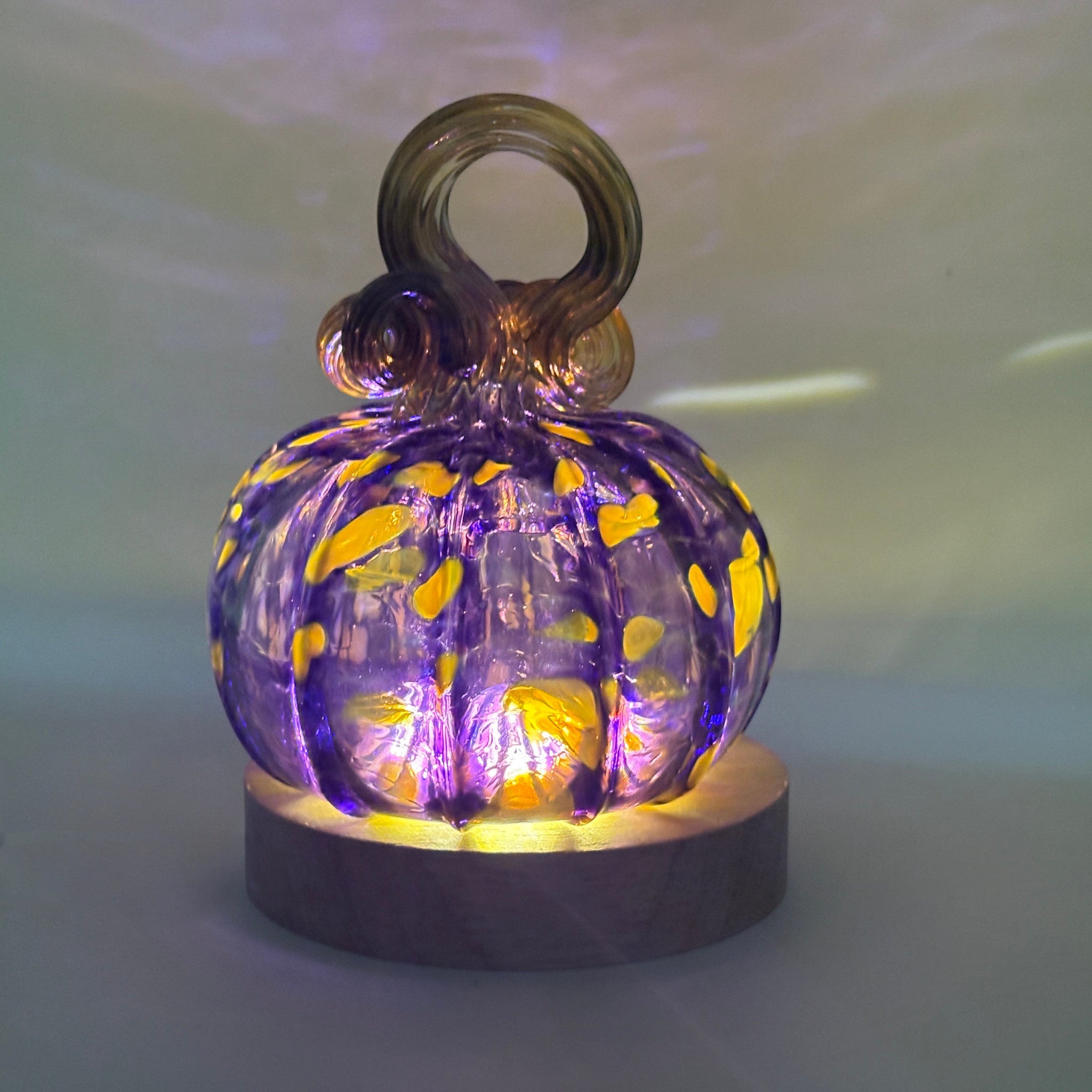 A handcrafted glass pumpkin with a loop stem, illuminated from within by an LED display light with a round wooden base.  