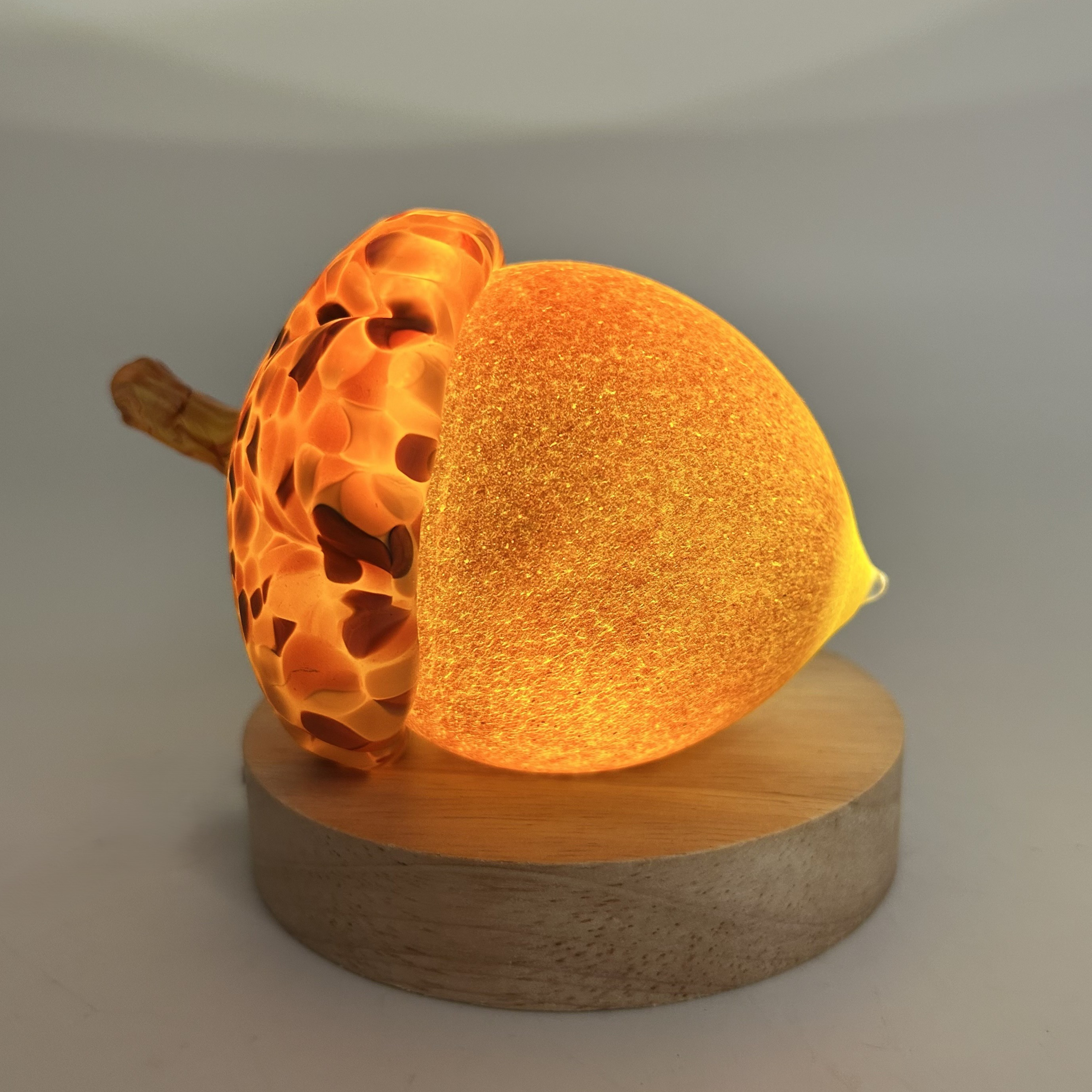 A handcrafted glass acorn with a honeycomb-patterned cap and a small brown stem, displayed on a round wooden LED light display base against a neutral gray background.