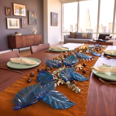 A styled dining table in a modern living room setting, featuring a rust-orange runner, blue glass leaf decorations, and green plates with cream napkins.