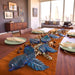 A styled dining table in a modern living room setting, featuring a rust-orange runner, blue glass leaf decorations, and green plates with cream napkins.
