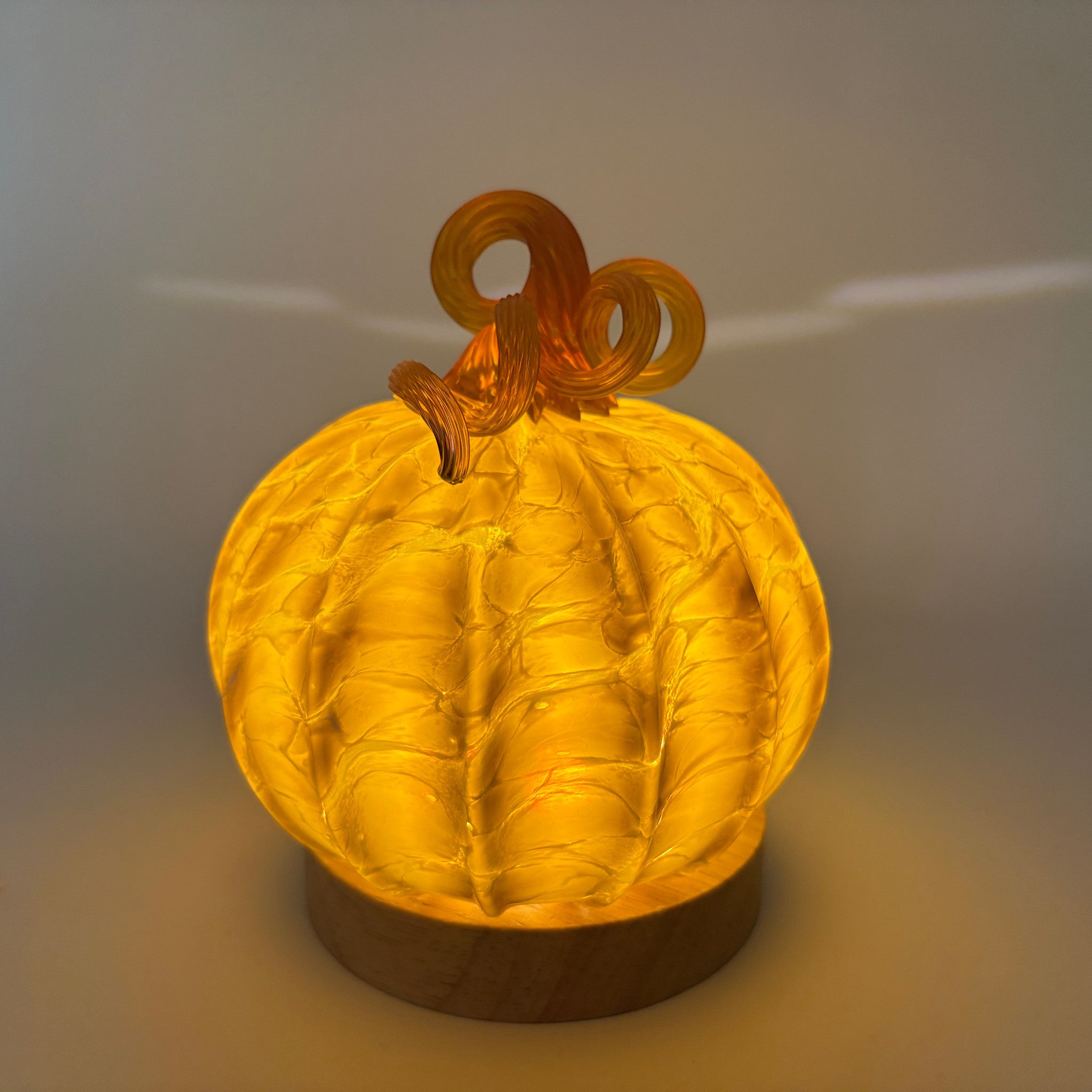 A glowing hand-crafted glass pumpkin  with a textured ribbed body and warm amber stem, displayed on an LED light with a wooden base.  