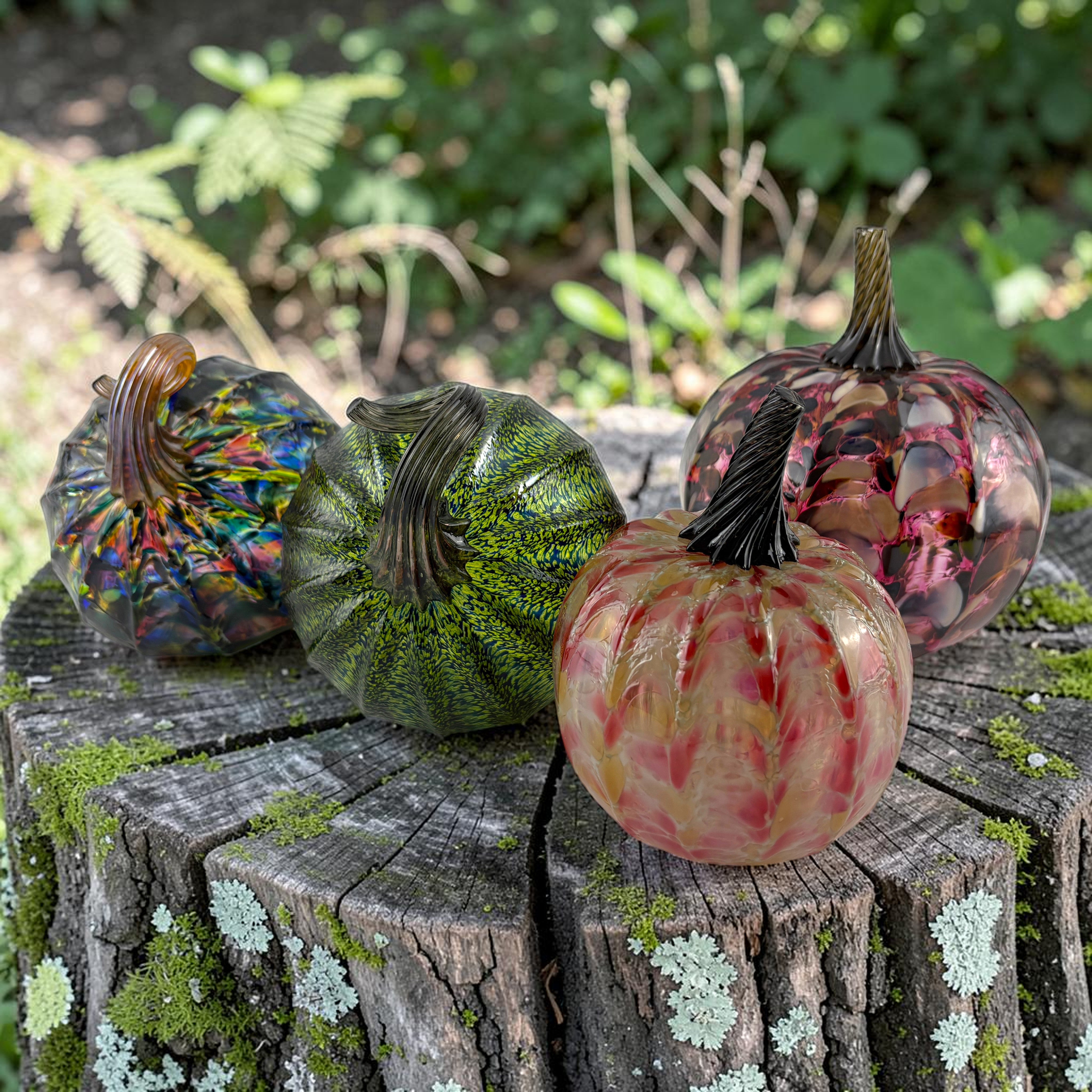 Four decorative glass pumpkins with textured, marbled patterns in green, red, and blue, displayed on a mossy tree stump in an outdoor garden setting.