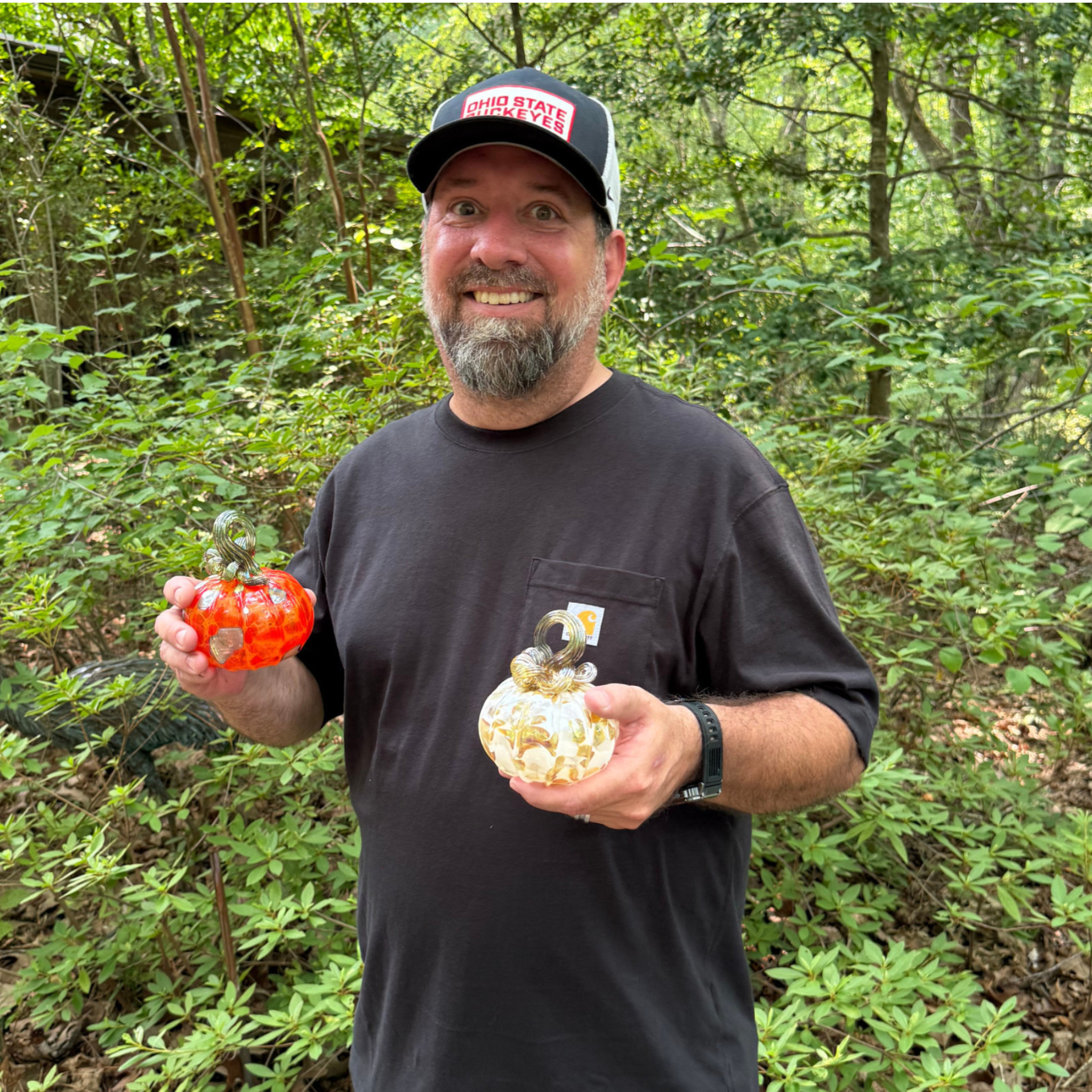 A smiling man wearing a black t-shirt and a black baseball cap, holding two decorative glass pumpkins in a lush green forest setting.