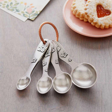 Set of four pewter measuring spoons with floral designs on a wooden surface, next to a plate of cookies.