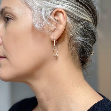 A close-up side profile of a woman with silver-gray hair wearing a delicate gold hoop earring with a small dangling bead, against a soft neutral background.