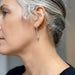 A close-up side profile of a woman with silver-gray hair wearing a delicate gold hoop earring with a small dangling bead, against a soft neutral background.