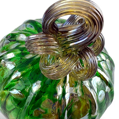 A close-up of a handcrafted glass pumpkin, featuring a textured green glass body with amber and gold marbling, topped with a large coiled gold glass stem.   