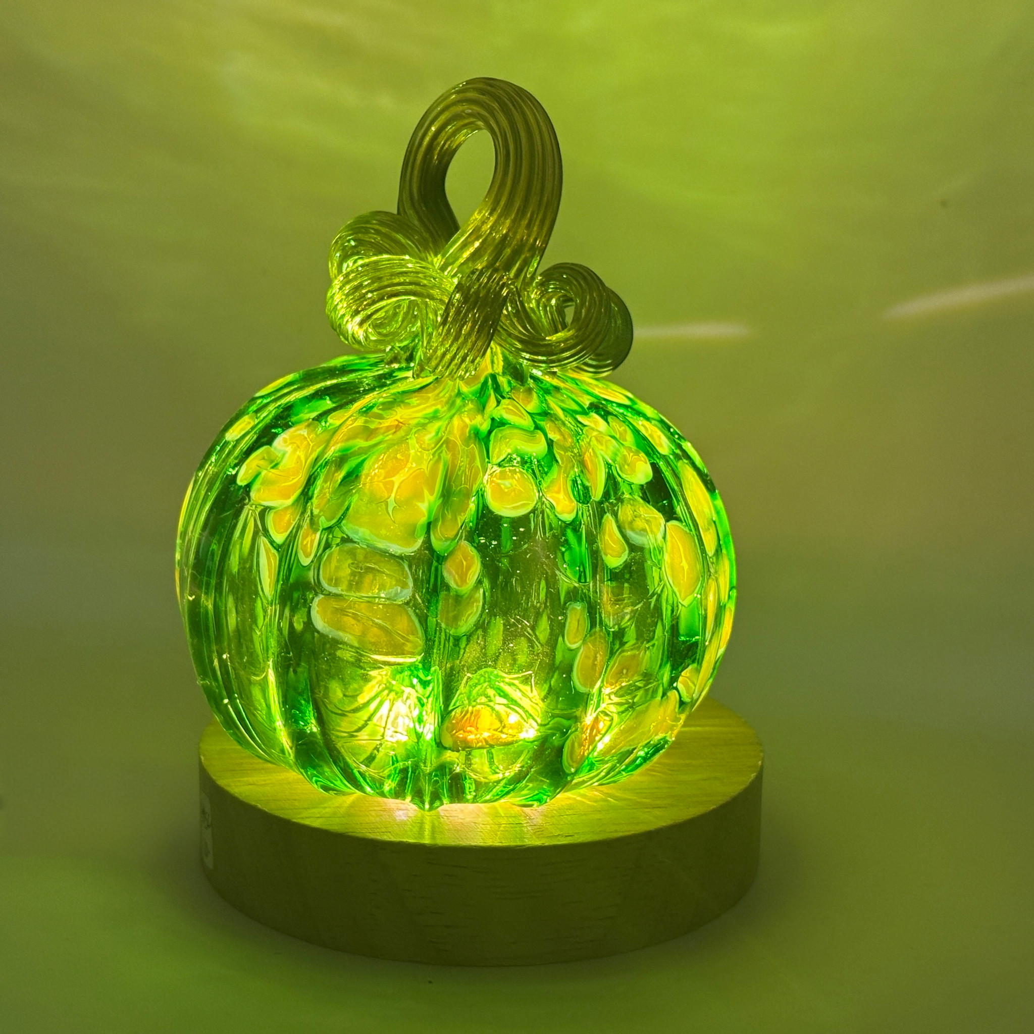 A glowing green glass pumpkin with a twisted stem, illuminated from within by an LED light with a round wooden base to cast a warm yellow light against a green-tinted background.