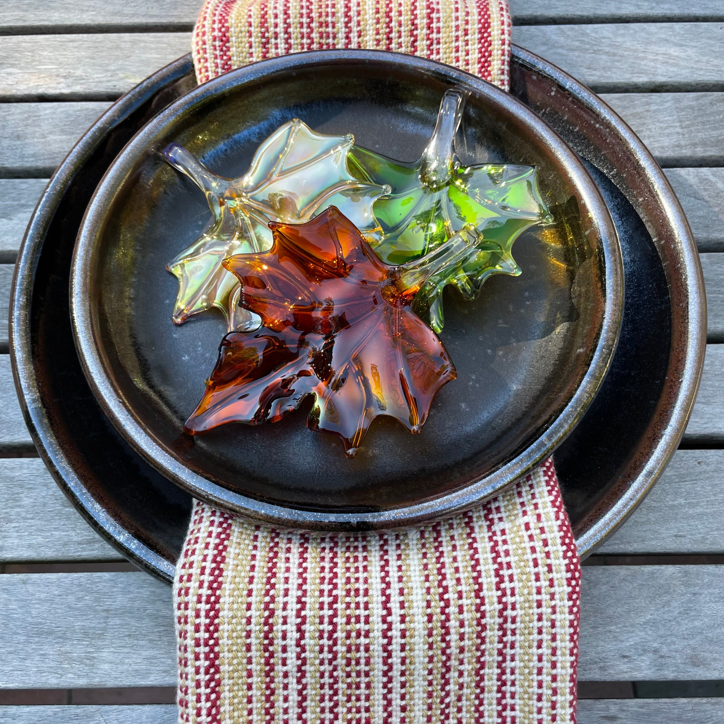 Three hand-blown glass maple leaves in amber, green, and gold tones, arranged on a dark ceramic plate with a striped napkin on a weathered wooden table.