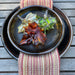 Three hand-blown glass maple leaves in amber, green, and gold tones, arranged on a dark ceramic plate with a striped napkin on a weathered wooden table.
