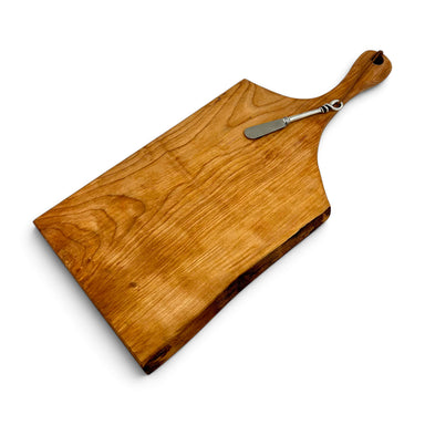 A free-form wooden serving board with a live edge and a rounded handle, shown with a small pewter pate utensil resting on top.