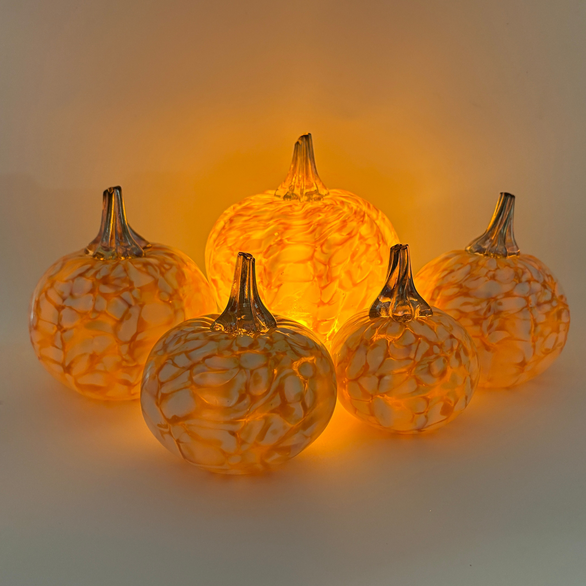 Five decorative glass pumpkins arranged together, glowing warmly from an internal light. The pumpkins have a textured, marbled pattern in orange and cream, with short brown stems, set against a plain light background.