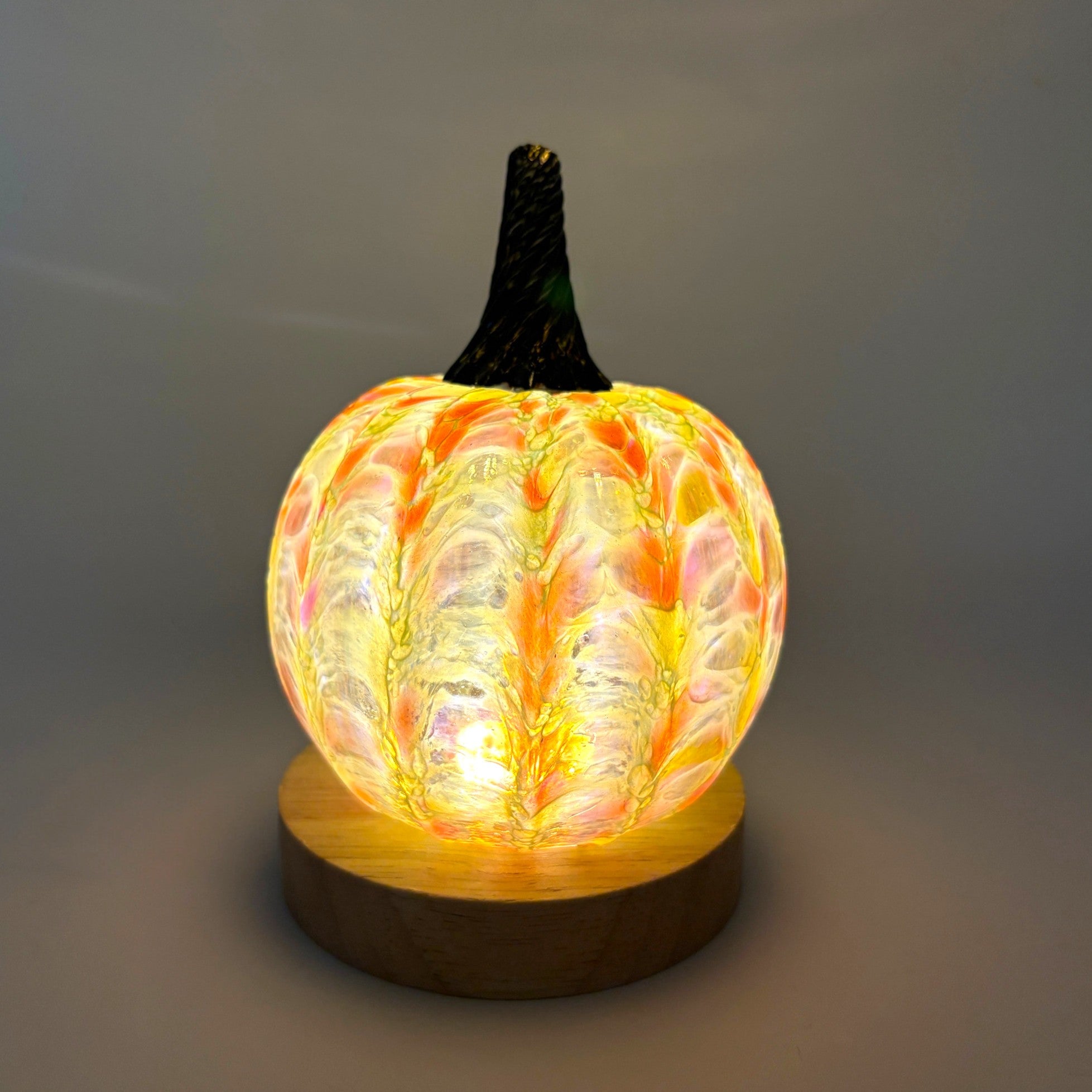 A handcrafted glass pumpkin with a marbled glass body in yellow, orange, and white, topped with a dark stem and resting on an LED display light with a round wooden base.  