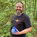 A smiling man with a beard stands outdoors in a wooded setting, holding two decorative glass ornaments. One ornament is green with yellow floral detailing, and the other is blue with white speckling.