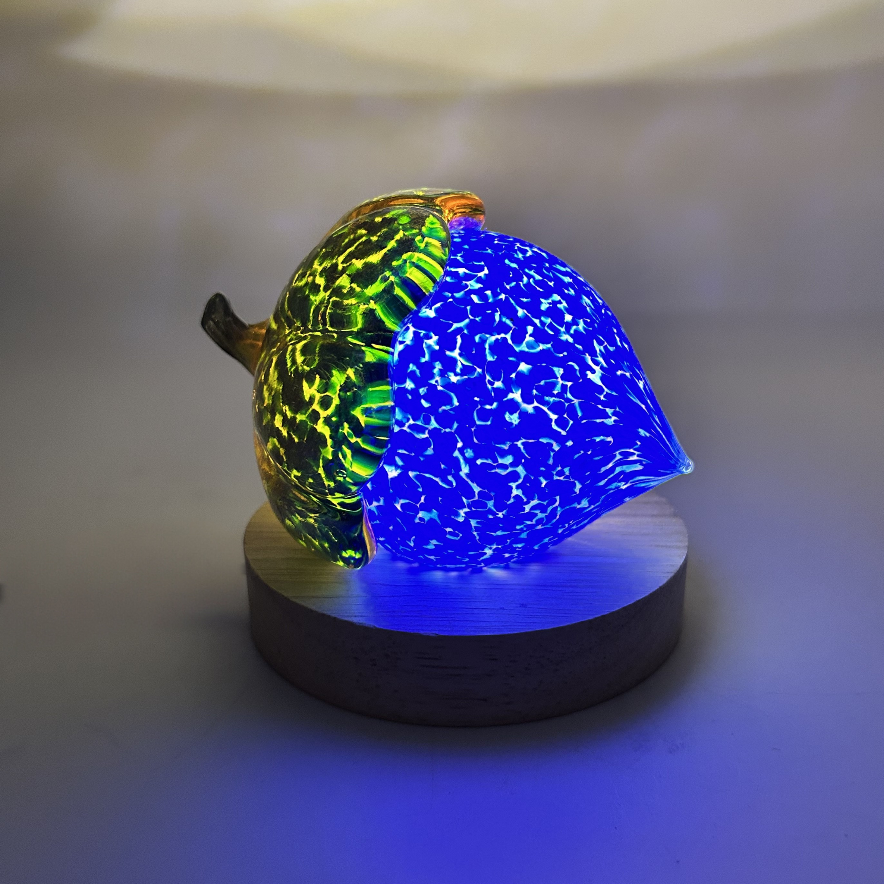 A decorative glass acorn sculpture on a round wooden base, lit to glow with a bright blue interior and a green-yellow patterned exterior, set against a softly lit neutral background.
