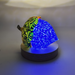 A decorative glass acorn sculpture on a round wooden base, lit to glow with a bright blue interior and a green-yellow patterned exterior, set against a softly lit neutral background.