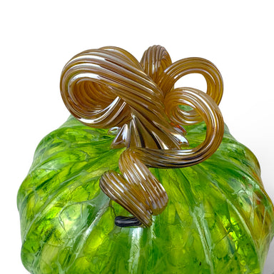A close-up of a hand-blown glass pumpkin with a ribbed green body and a thick, glossy amber-brown stem.   