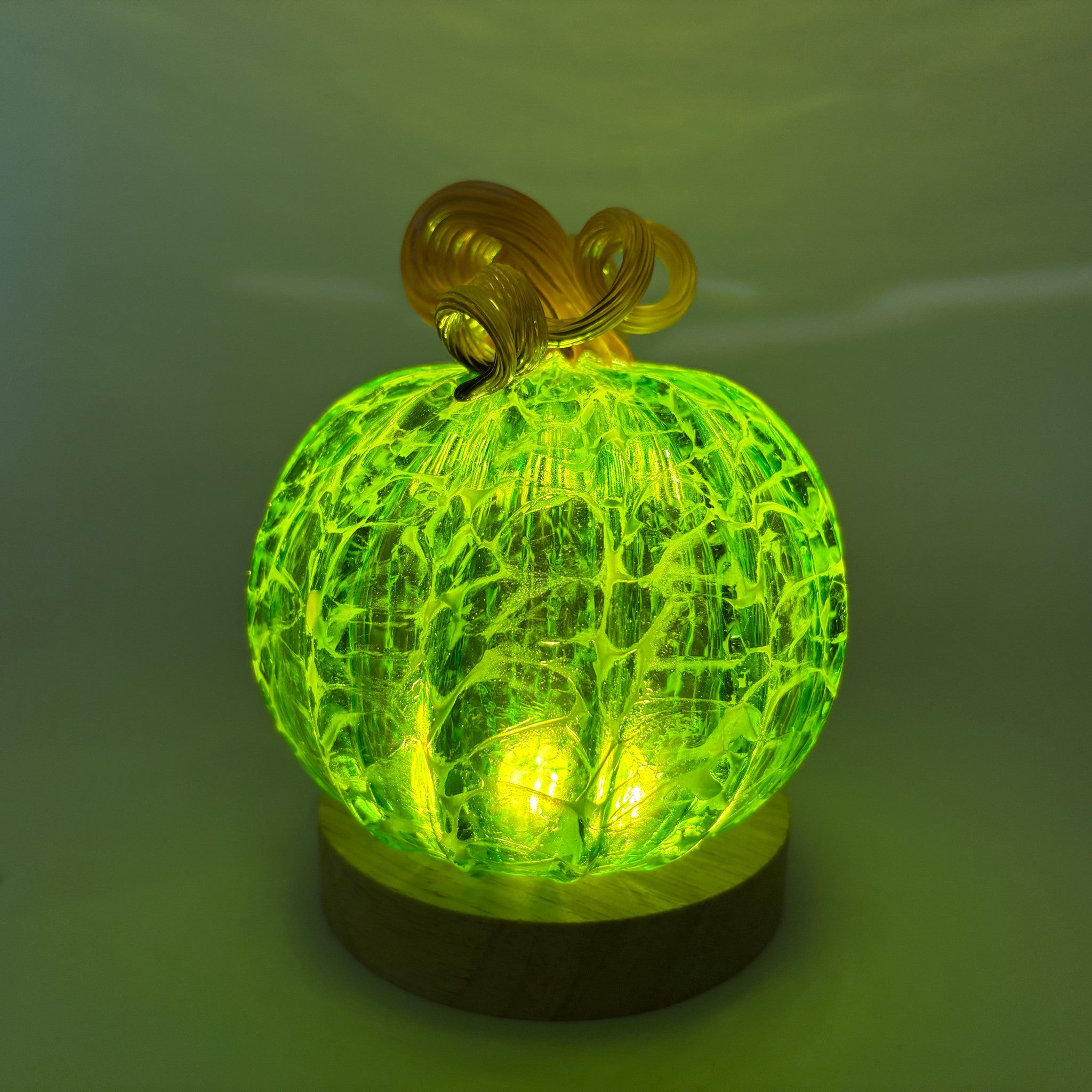 A glowing decorative pumpkin with a textured green cracked-glass sphere, a brown stem, displayed on and LED light with a round wooden base.    