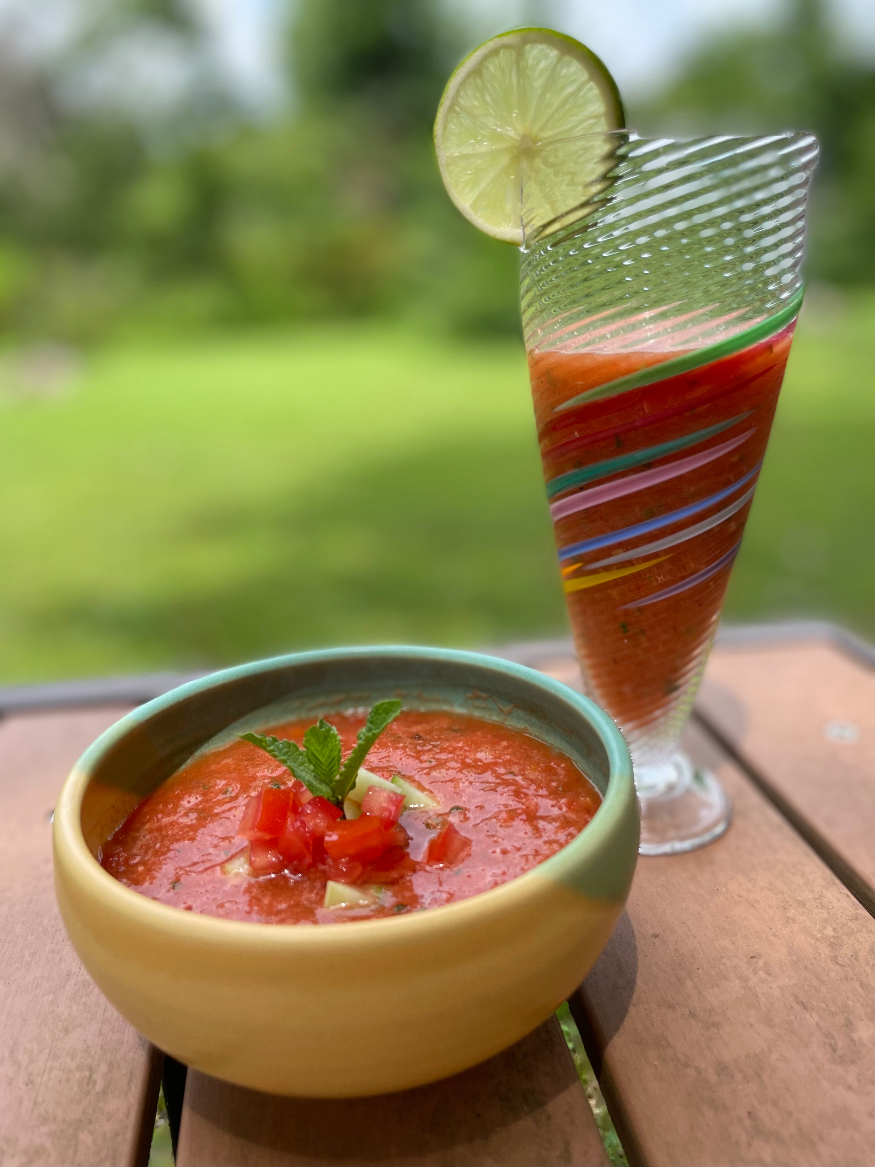 Bowl of gazpacho with a colorful glass of tomato soup on a wooden surface outdoors.