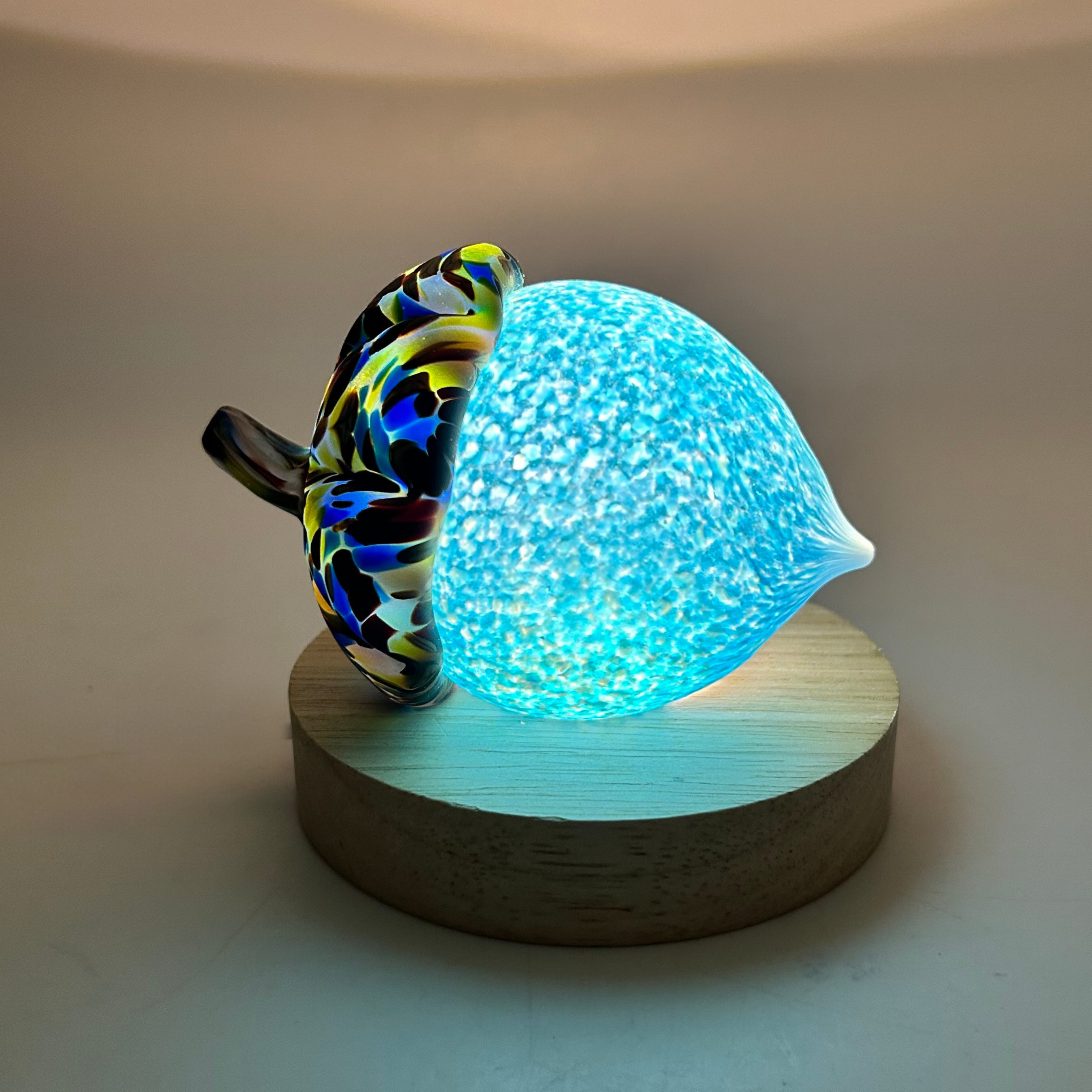 A decorative acorn with a glowing cerulean blue body and a multicolored patterned cap, displayed on a round wooden LED light base. 