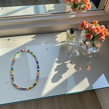 Colorful beaded necklace on a glass surface with a vase of flowers and a bottle of perfume.