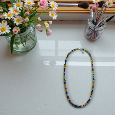 Colorful beaded necklace on a white surface with flowers and makeup brushes in the background