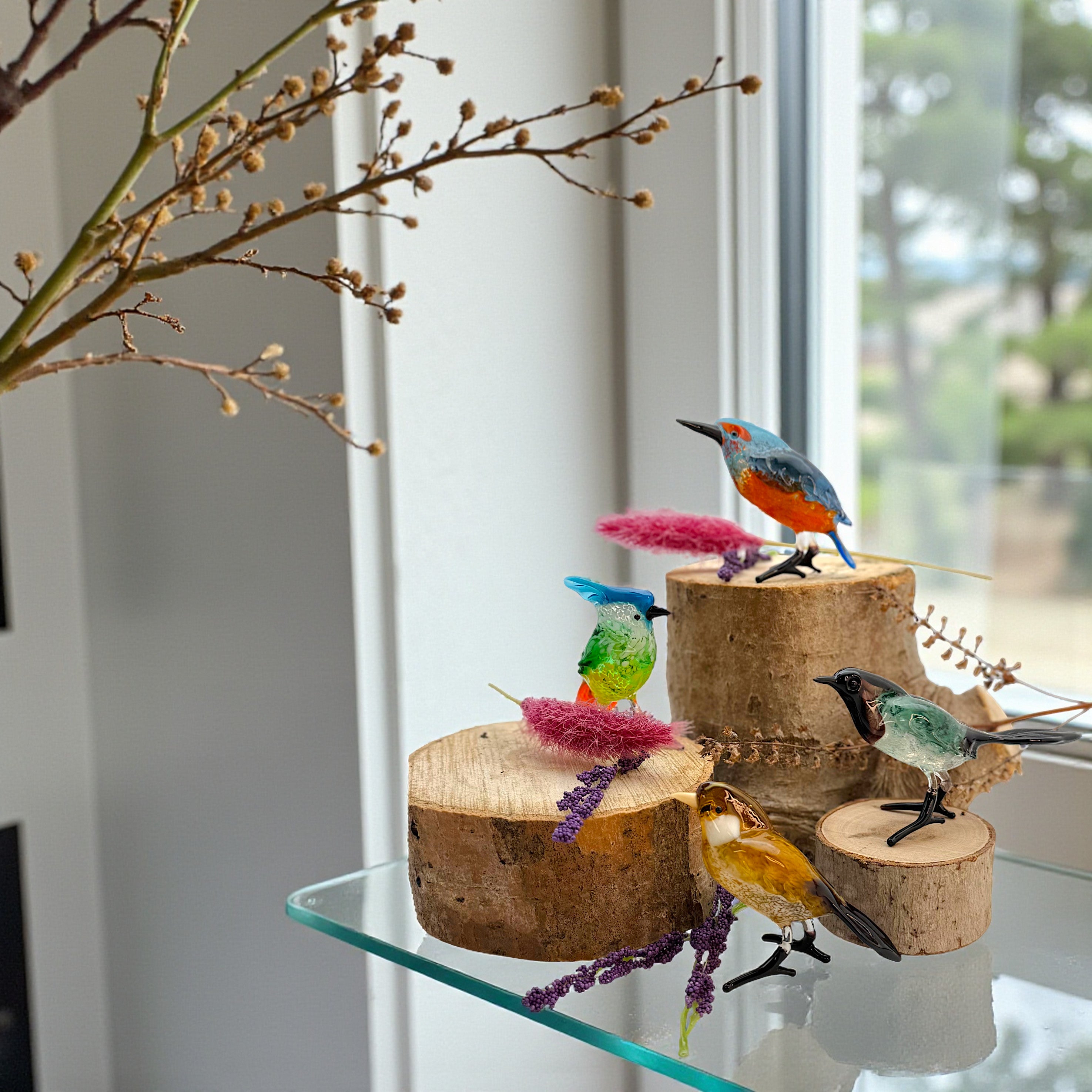 Colorful bird figurines on wooden perches on a glass shelf with branches in the background.