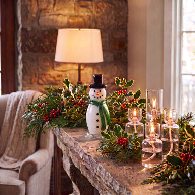 A handcrafted glass snowman figurine with a green scarf and black top hat sits on a stone fireplace mantel, surrounded by holiday greenery with red berries and lit glass candle holders in a cozy indoor setting.