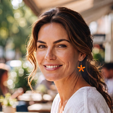 A smiling woman with brown hair wearing a white top and an orange flower-shaped dangle earring, photographed in a softly blurred outdoor cafe setting.