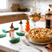 A styled kitchen countertop with a baked pie on a plate in the foreground, decorative handcrafted glass snowman and pumpkin figures, green handcrafted glass leaf sculptures, and a glass jar of Halloween cookies. 