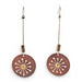A pair of dangling earrings with circular sunburst-patterned wooden discs in reddish-brown and black, accented with small white dots and a gold center, hanging from silver-toned wires on a white background.