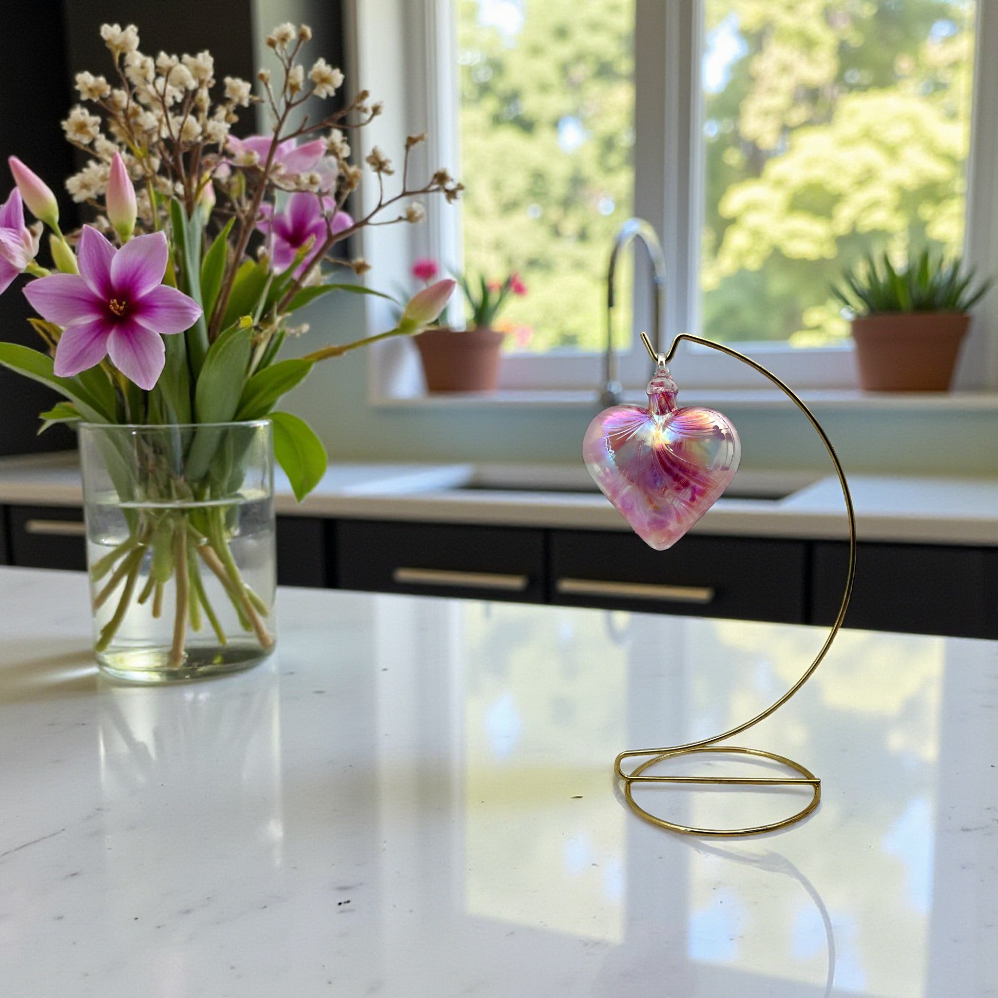 Decorative heart-shaped hand-blown glass ornament on a stand next to a vase of flowers in a kitchen.
