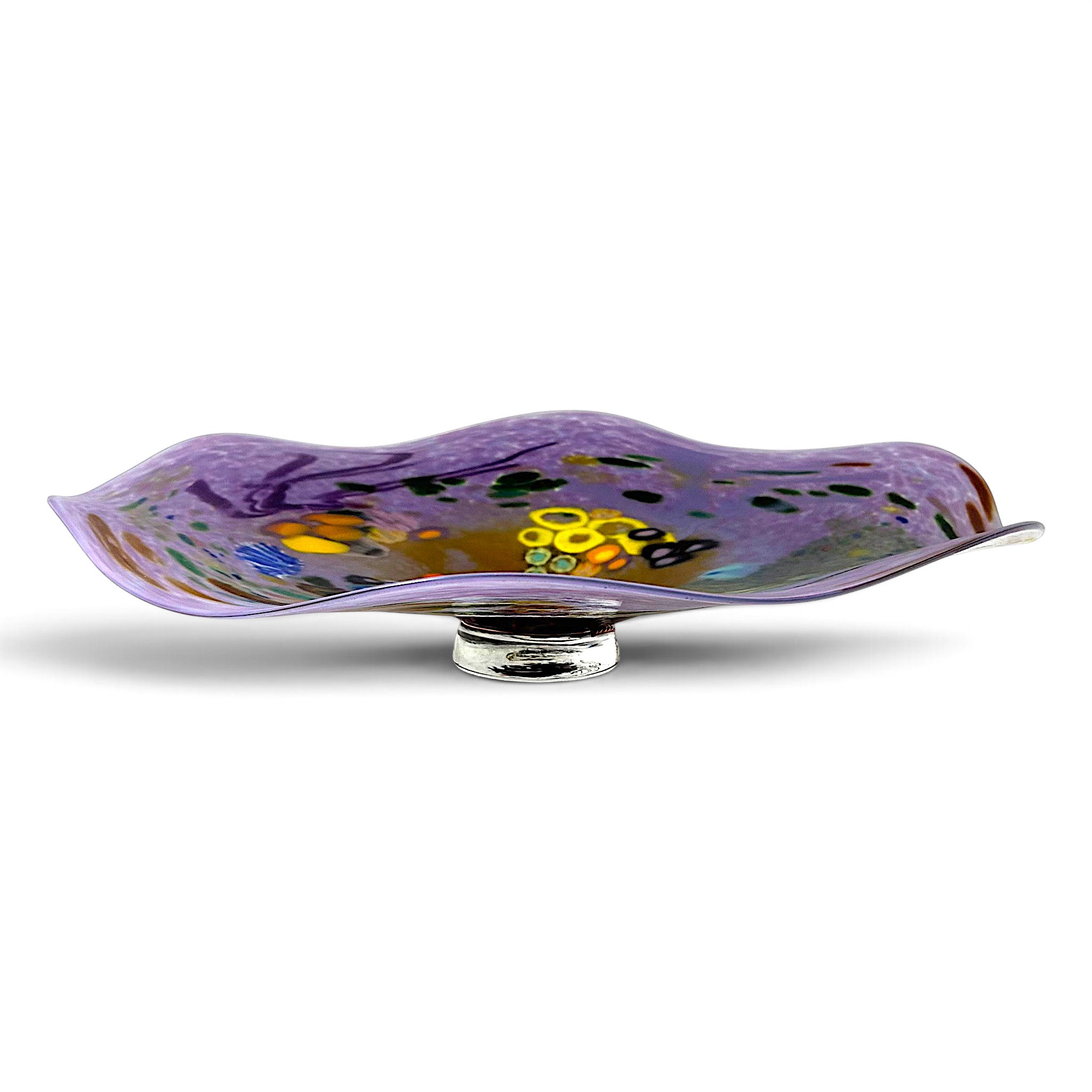 A decorative hand-blown glass reef bowl with a wavy, open top design, featuring a purple and gold pattern with small multicolored accents, incorporating a glass foot for using a a statement centerpiece on a table.