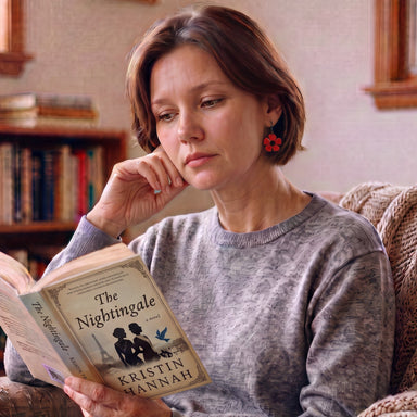 A woman with short brown hair sits in a cozy armchair, holding and reading the book 'The Nightingale' by Kristin Hannah. She is wearing a textured gray knit sweater and red flower-shaped earrings, with a bookshelf in the background.