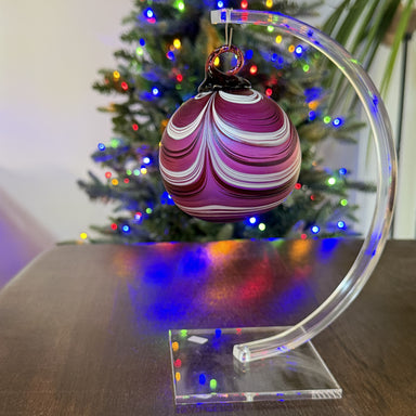 Decorative Christmas ornament on a clear stand with a Christmas tree in the background.