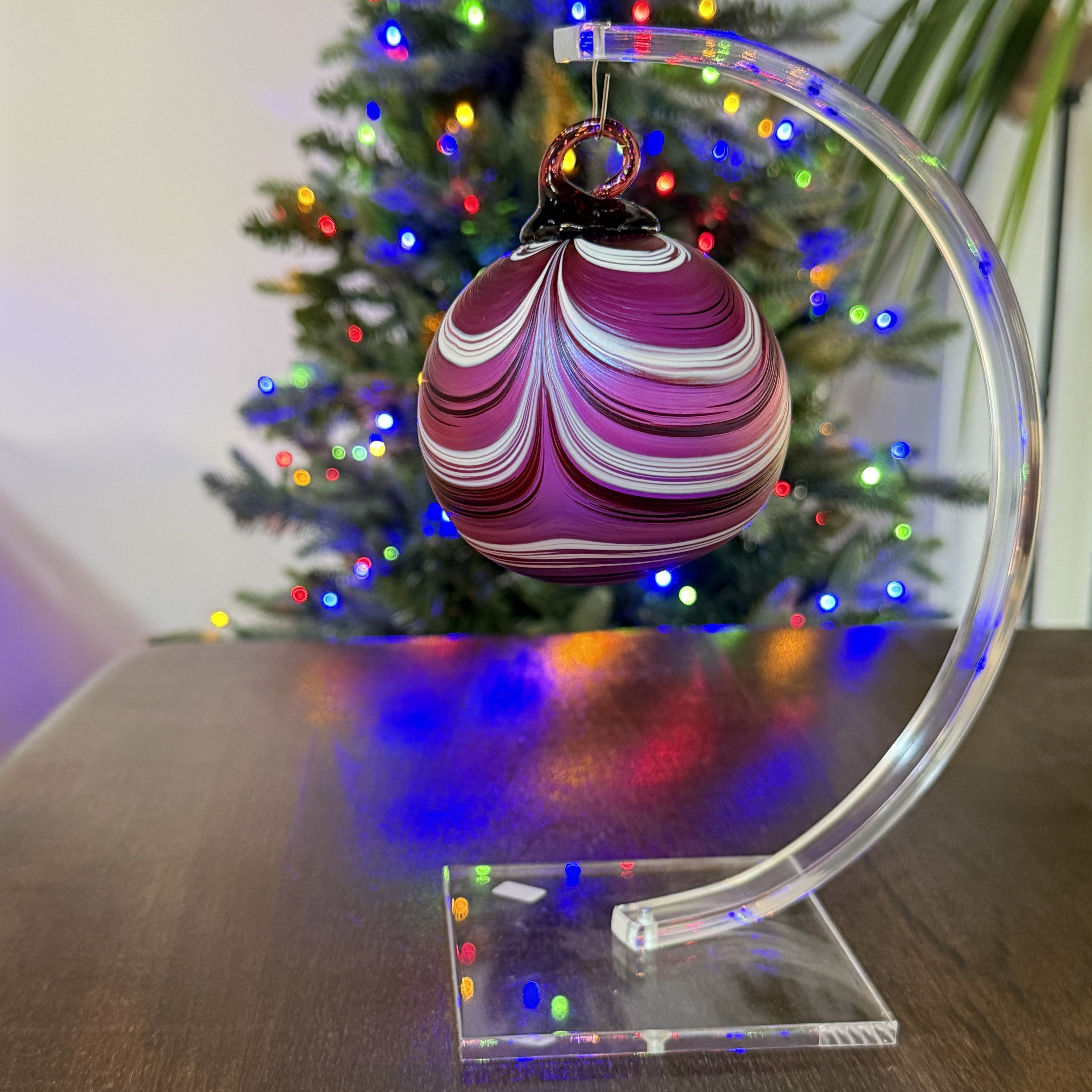 Decorative Christmas ornament on a clear stand with a Christmas tree in the background.