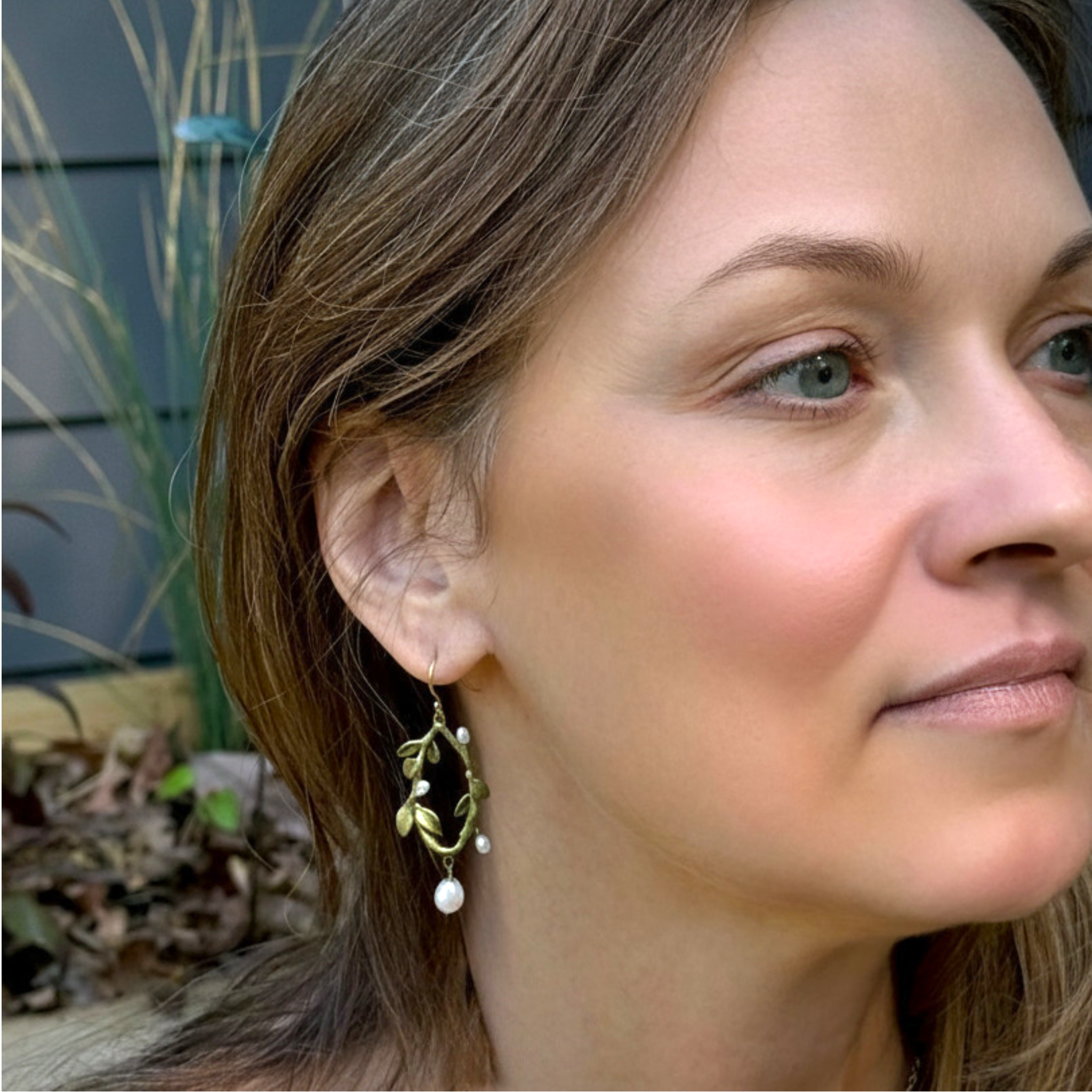 A close-up lifestyle shot of a woman wearing a dangling earring. The earring features an olive-green leafy vine design with small white pearl-like beads and a white teardrop charm.