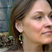 A close-up lifestyle shot of a woman wearing a dangling earring. The earring features an olive-green leafy vine design with small white pearl-like beads and a white teardrop charm.