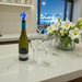 Wine bottle with a blue stopper, wine glasses, and flowers on a kitchen counter with city lights in the background.