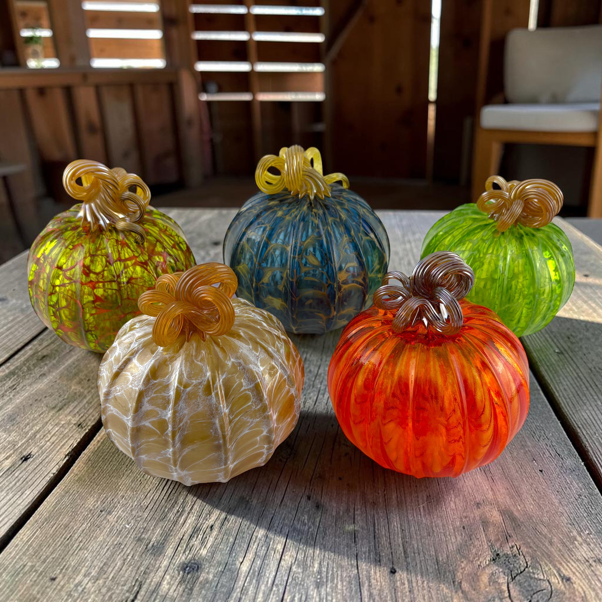 Five decorative glass pumpkins in autumn colors arranged on a rustic wooden table. The pumpkins include orange, yellow, green, blue, and cream tones with textured marbled patterns and twisted stem details.