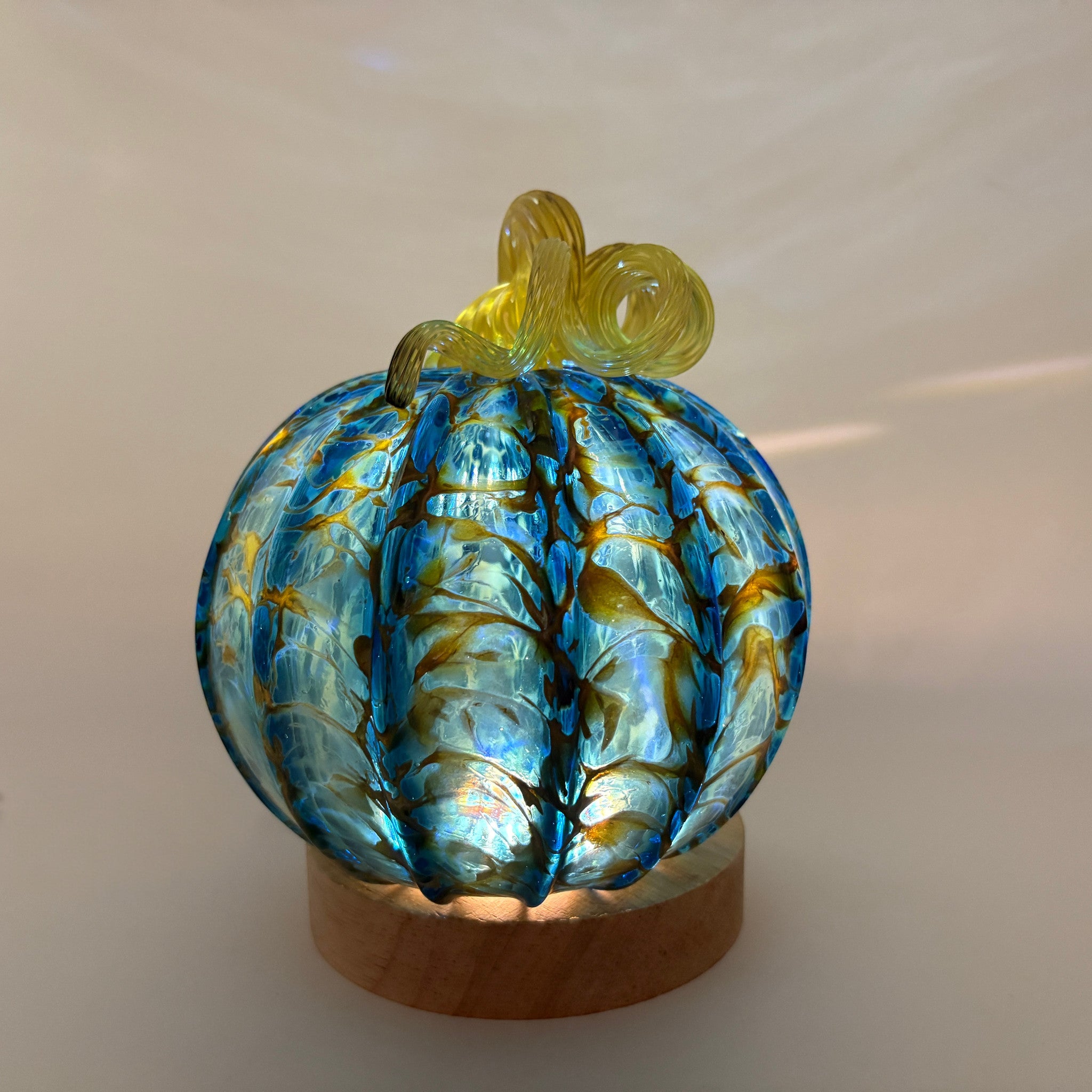 A hand-blown glass decorative pumpkin with a blue and yellow-gold marbled pattern, resting on a small wooden base display light. 