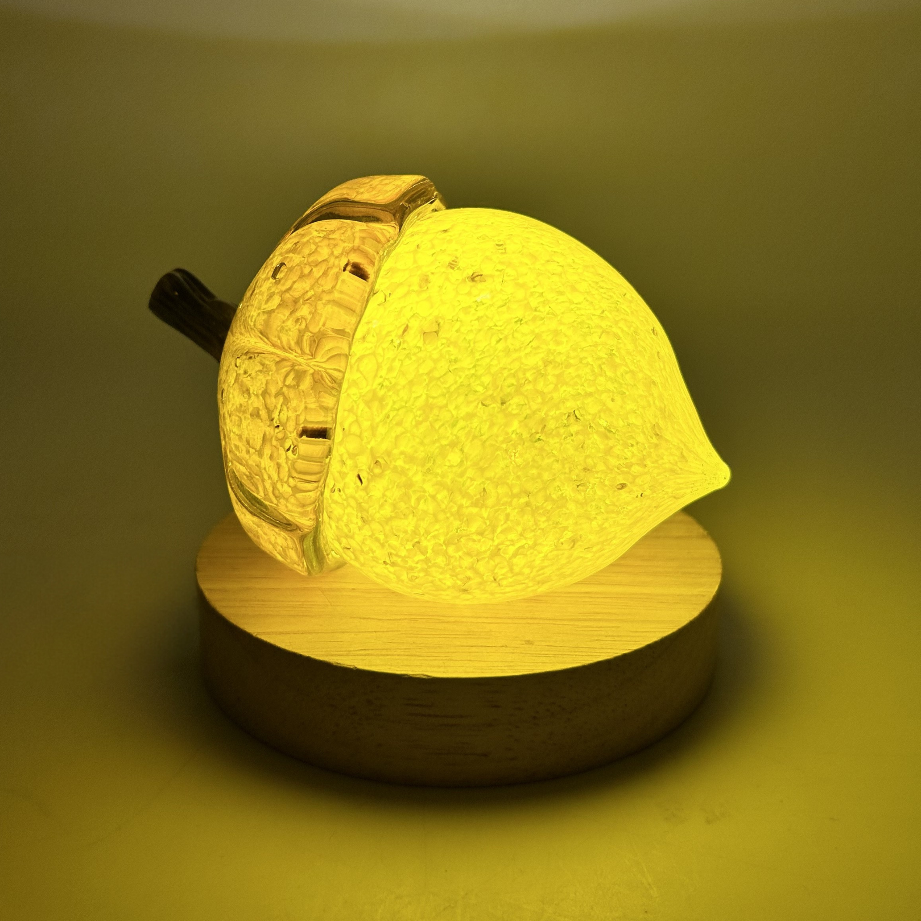 A glowing acorn with a textured yellow body, a dark brown stem, on a round wooden base LED display light, photographed against a warm yellow background.