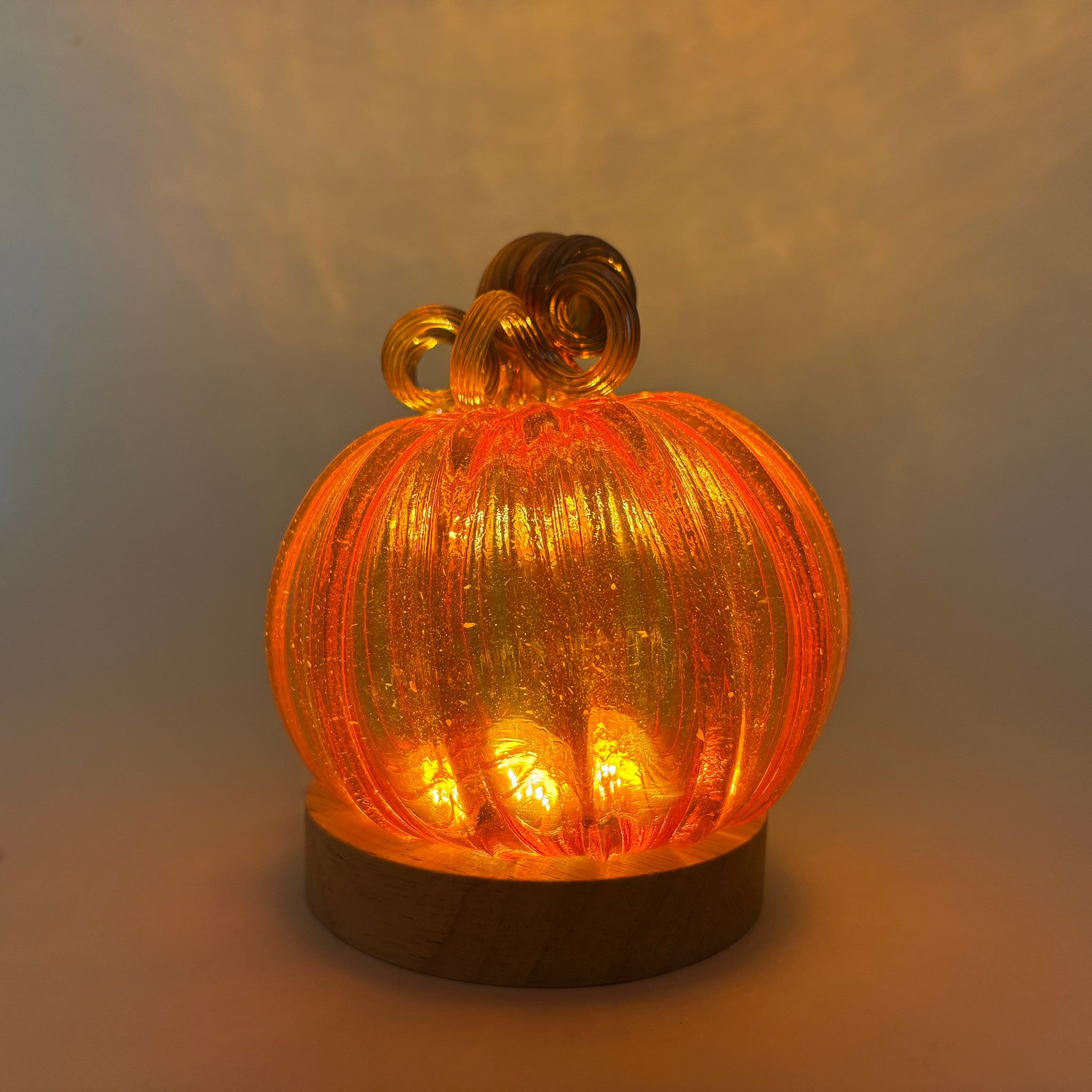 An illuminated handcrafted orange glass pumpkin, displayed on an LED light with a wooden base.  